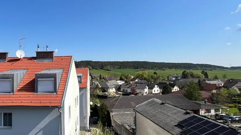 Blick über Dächer und grüne Hügel einer ländlichen Umgebung unter blauem Himmel.