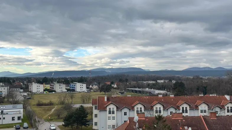 Weitläufiger Blick über eine Wohnsiedlung mit Häusern, Bäumen und Bergen im Hintergrund unter bewölktem Himmel.