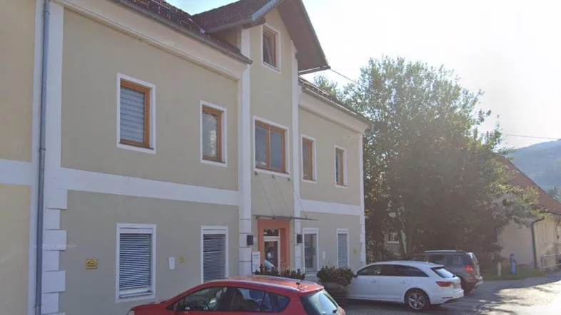 Ansicht eines Mehrfamilienhauses mit beiger Fassade, braunen Fenstern und geparkten Autos auf der Straße.