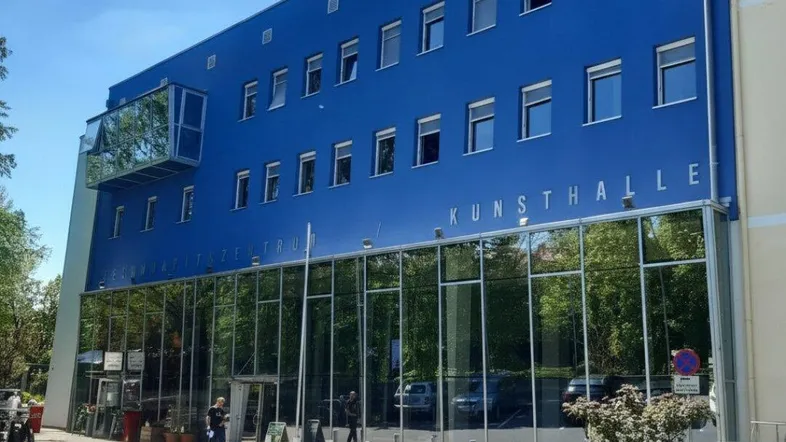 Modernes blaues Gebäude mit großer Glasfassade und vielen Fenstern unter strahlend blauem Himmel.