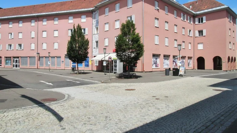 Mehrstöckiges Gebäude mit lachsfarbener Fassade und Geschäften im Erdgeschoss an einem sonnigen Platz.