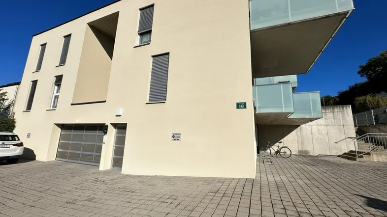 Moderne Gebäudefassade mit hellbeigem Putz, großen Garagentoren und verglasten Balkonen unter blauem Himmel.