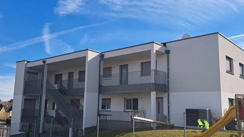 Modernes Mehrfamilienhaus mit Balkonen und einem kleinen Spielplatz im Vordergrund unter blauem Himmel.
