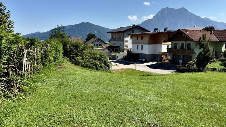 Grüne Wiese mit Blick auf umliegende Häuser und majestätische Berge im Hintergrund unter blauem Himmel.