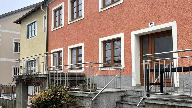 Vorderansicht eines Wohnhauses mit orangeroter Fassade, braunen Fenstern und einem Eingangsbereich mit Treppe.