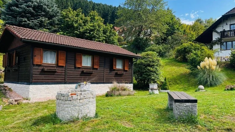 Kleines Holzhaus mit Gartenbereich, Sitzbank und dekorativem Brunnen in ländlicher Umgebung.