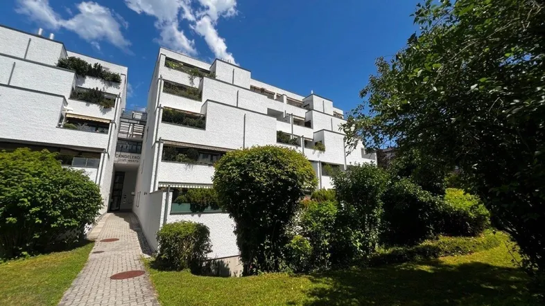 Modernes Mehrfamilienhaus mit weißer Fassade, Balkonen und gepflegter Grünanlage unter blauem Himmel.