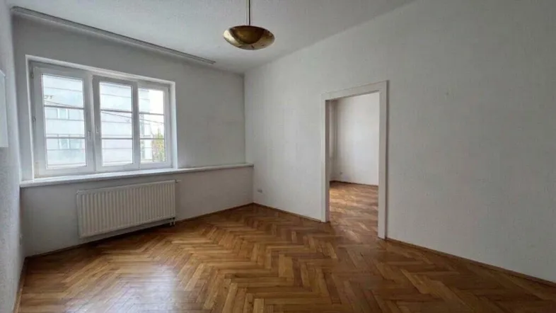 Helles Zimmer mit Fischgrätparkettboden und Fenster, das zu einem weiteren Raum führt.