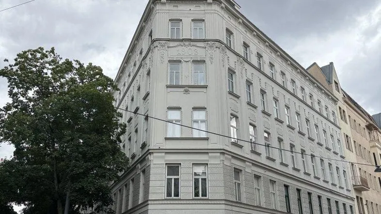 Eckgebäude mit klassischer Architektur und vielen Fenstern an einer städtischen Straße unter bewölktem Himmel.