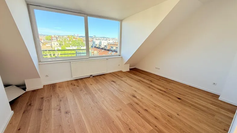 Heller Raum mit Holzboden, Dachschrägen und großem Fenster mit Stadtblick.