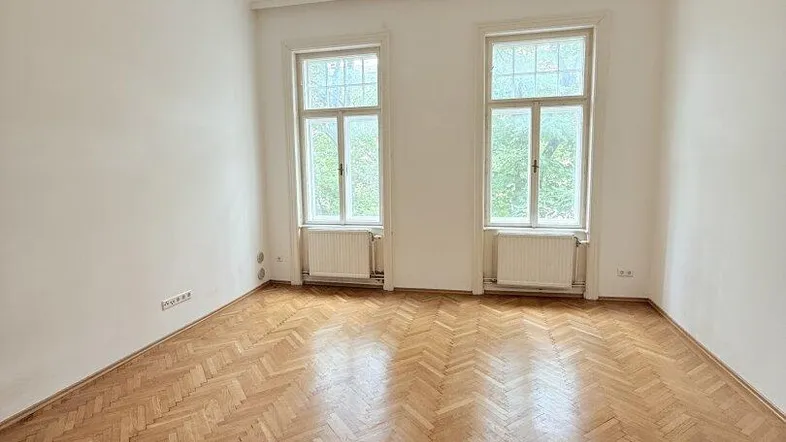 Helles Zimmer mit Fischgrätparkett, hohen Decken und Stuckverzierung an der Decke.