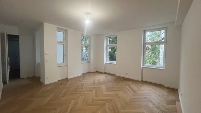 Helles Wohnzimmer mit Fischgrätparkett und mehreren Fenstern, das viel Tageslicht hereinlässt.