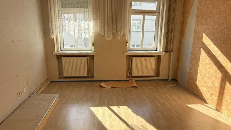 Helles Zimmer mit Parkettboden, Deckenventilator und zwei Fenstern, bereit zur Renovierung.