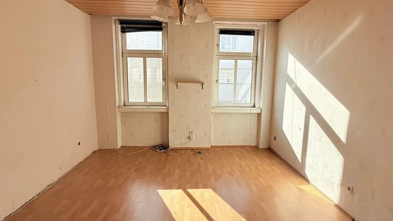 Helles Zimmer mit Holzdeckenverkleidung, Laminatboden und zwei Fenstern, die viel Sonnenlicht hereinlassen.