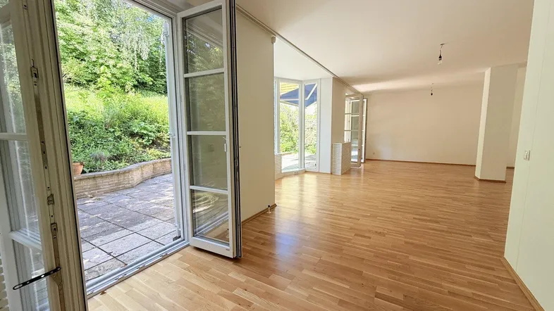 Helles, leeres Zimmer mit Holzboden und großen Fenstern, die Zugang zu einer Terrasse bieten.
