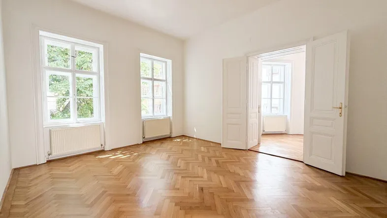 Helles Zimmer mit Fischgrätparkett, weißen Wänden und hohen Doppeltüren, die zu einem weiteren Raum führen.