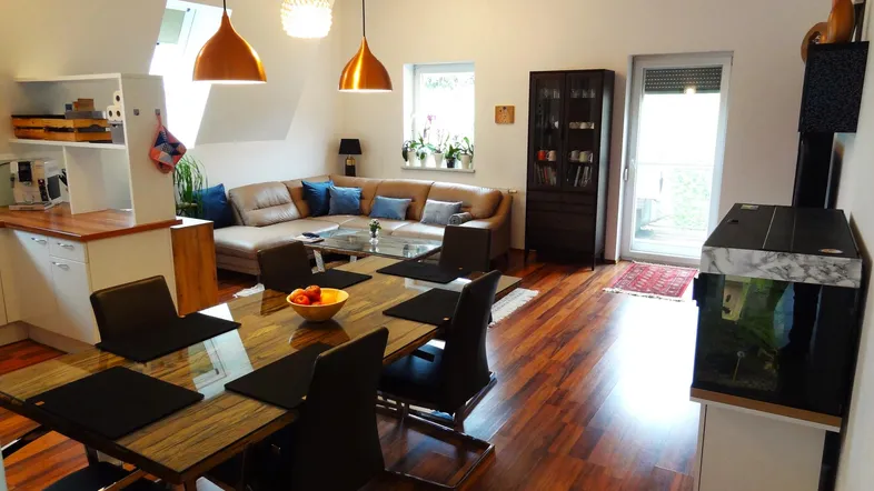 Geräumiger offener Wohn- und Essbereich mit Holzboden, beiger Eckcouch und Zugang zum Balkon.