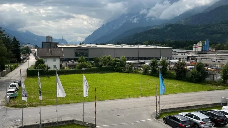 Blick auf eine grüne Wiese mit umliegenden Industriegebäuden und majestätischen Bergen im Hintergrund unter bewölktem Himmel.