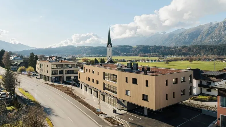 Moderne Wohngebäude in ländlicher Umgebung mit Bergblick und Kirchturm im Hintergrund.