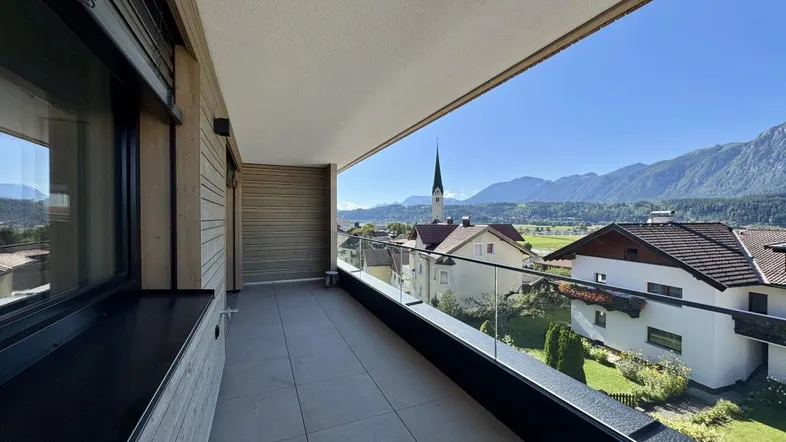 Balkon mit grauen Fliesen, Glasgeländer und weitem Alpenblick bei Sonnenschein.