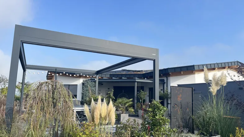 Moderne Hausfassade mit Pergola, üppigem Garten und gemütlichem Außenbereich unter blauem Himmel.