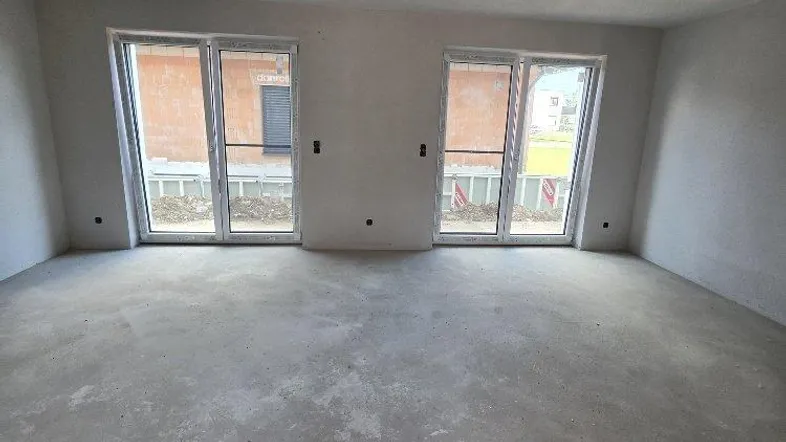 Unfertiger Innenraum mit Rohbauwänden, Betonboden und zwei großen Fenstertüren, die auf eine Baustelle blicken.