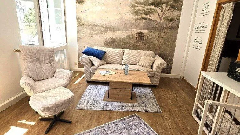 Helles Wohnzimmer mit Holzboden, bequemen Möbeln und einer dekorativen Wandtapete mit Landschaftsmotiv.