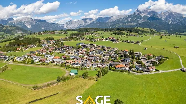 Weitläufiger Panoramablick auf ein idyllisches Dorf mit grünen Wiesen und majestätischen Bergen.