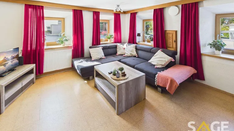 Gemütliches Wohnzimmer mit großem Ecksofa, Holzbalken und hellen Fenstern mit roten Vorhängen.