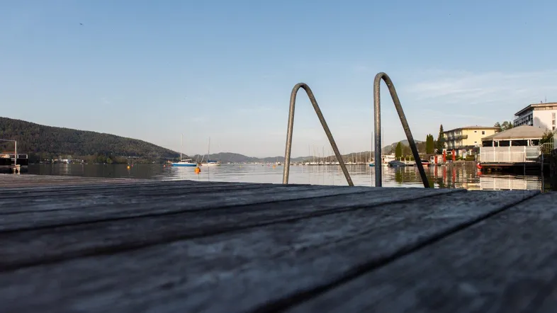 Holzsteg am Seeufer mit Blick auf die umliegende Berglandschaft und Segelboote.