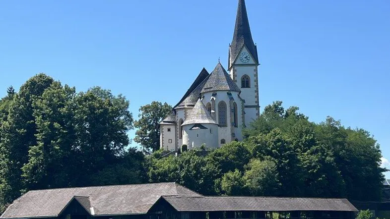 Historische Kirche am Seeufer mit traditionellen Bootshäusern und klarem blauen Himmel.