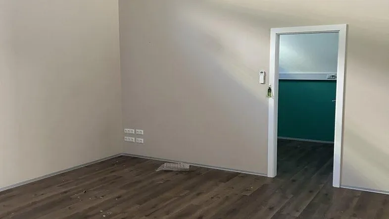 Leerer Büroraum mit Laminatboden, Klimaanlage und Zugang zu einem weiteren Zimmer mit farbiger Wand.
