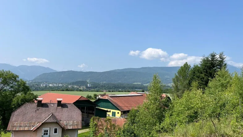 Ländliche Landschaft mit Häusern, grünen Feldern und Bergen im Hintergrund unter blauem Himmel.
