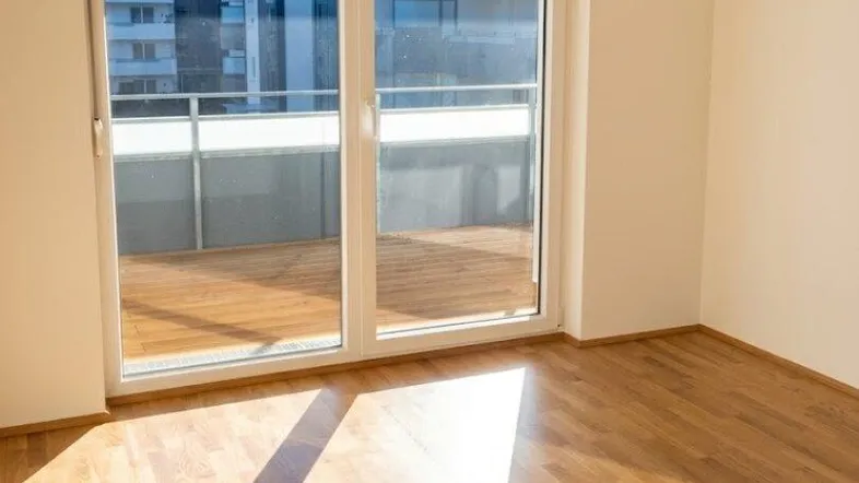 Helles Zimmer mit Holzboden und Zugang zu einem Balkon, mit Blick auf moderne Gebäude.