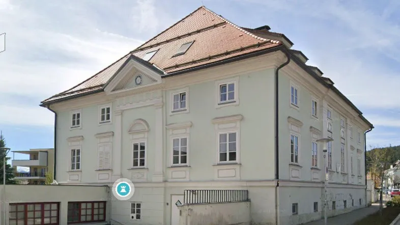 Historisches Gebäude mit klassischer Fassade, Satteldach und großen Fenstern in heller Farbgebung.