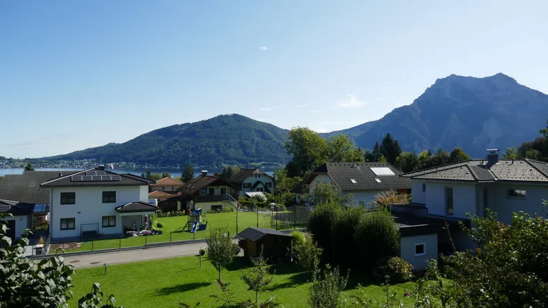 Panoramablick über eine Wohnsiedlung mit grünen Gärten, einem See und majestätischen Bergen im Hintergrund.