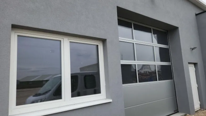Graue Fassade eines modernen Gebäudes mit einem Fenster und einem großen Sektionaltor, das Licht in den Innenraum lässt.