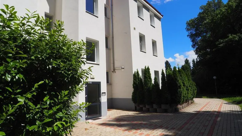 Weißes Mehrfamilienhaus mit gepflastertem Eingangsbereich und grünen Büschen unter blauem Himmel.