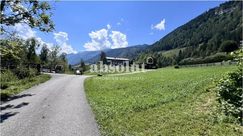 Landstraße führt durch grüne Wiesen mit Blick auf bewaldete Berge und ein entferntes Gebäude.