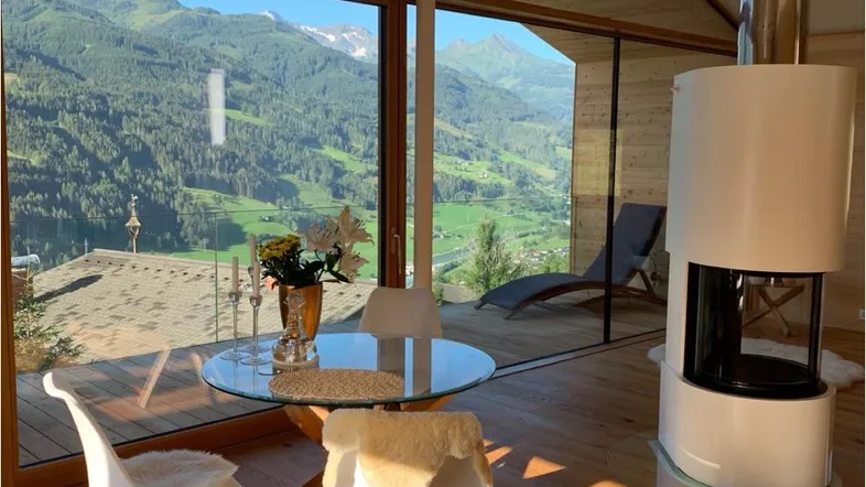 Helles Wohnzimmer mit Panoramablick auf die Berge, Holzboden und modernem Kaminofen.
