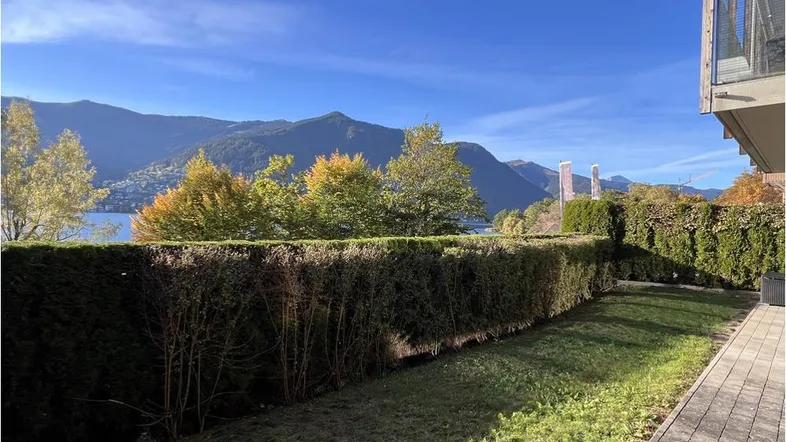 Gepflegter Gartenbereich mit Rasenfläche und Hecke, mit weitem Blick auf den See und die Berge.