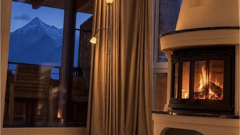 Gemütliches Wohnzimmer mit brennendem Kamin, großen Fenstern und beeindruckendem Bergblick am Abend.