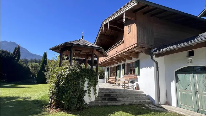 Traditionelles Haus im alpenländischen Stil mit großem Garten, Pavillon und Bergblick unter blauem Himmel.