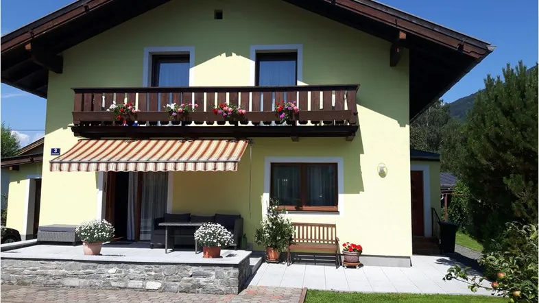 Sonnige Terrasse mit Markise und Balkon, umgeben von einem grünen Gartenbereich und Bergblick.