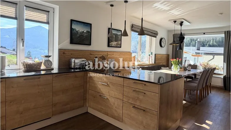 Moderne Küche mit Holzfronten, dunkler Arbeitsplatte und integriertem Essbereich mit Bergblick.