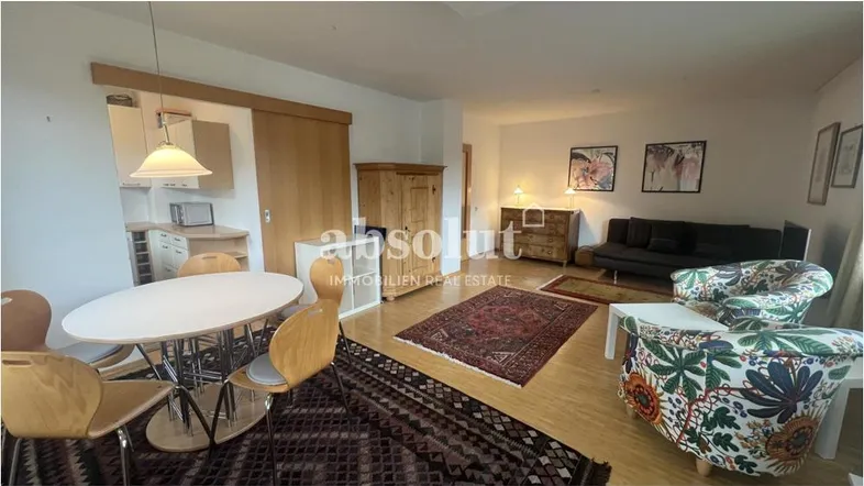 Gemütlicher Wohn-Essbereich mit Holzboden, Teppichen und einem Schrank aus hellem Holz, der den Raum strukturiert.