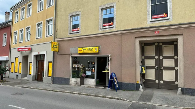 Fassade eines Mehrfamilienhauses mit Ladenlokalen im Erdgeschoss und Wohnungen darüber, an einer Straße gelegen.