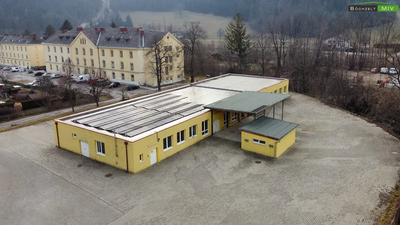 Luftaufnahme eines gelben Gebäudes mit Solarpaneelen auf dem Dach und einem größeren Gebäude im Hintergrund.
