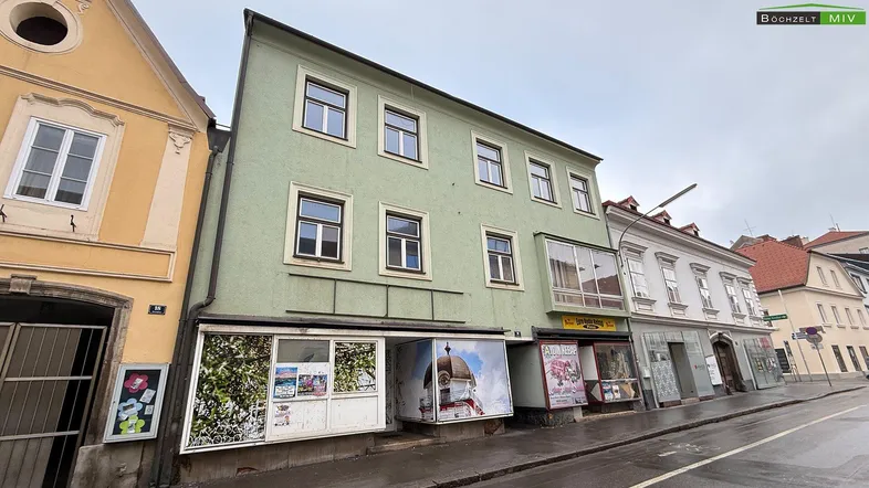 Grünes Mehrfamilienhaus mit Geschäften im Erdgeschoss und Wohnungen darüber an einer Stadtstraße.