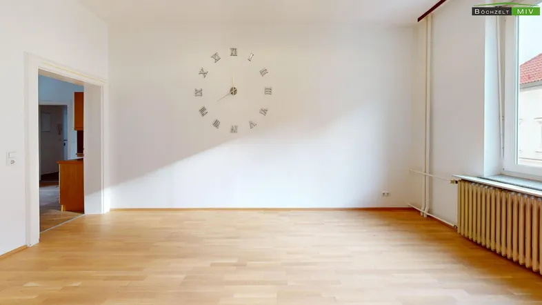 Helles Zimmer mit Parkettboden, großer Wanduhr und Heizkörper unter dem Fenster.